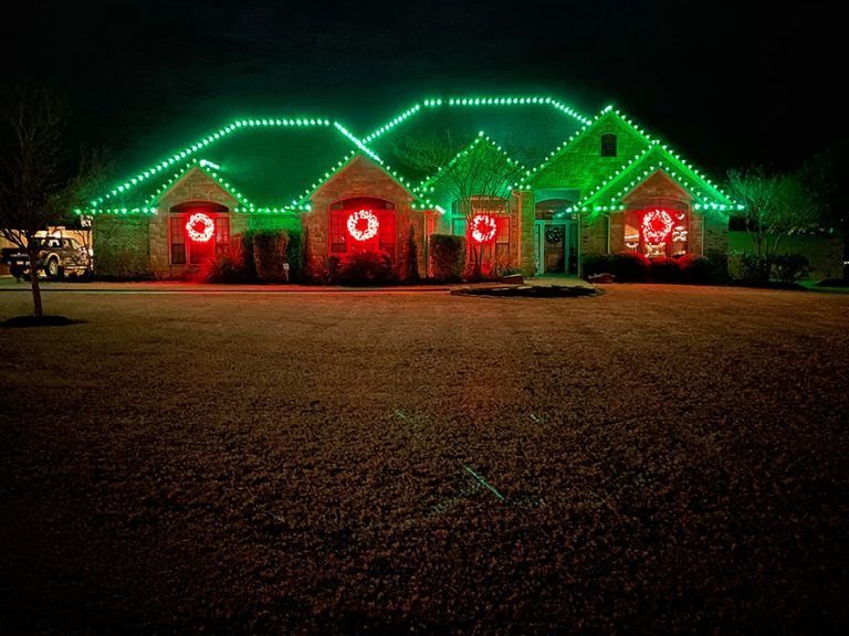 Green Christmas lights on the roofline and wreaths made of red Christmas lights in each window on a house in Leander TX.