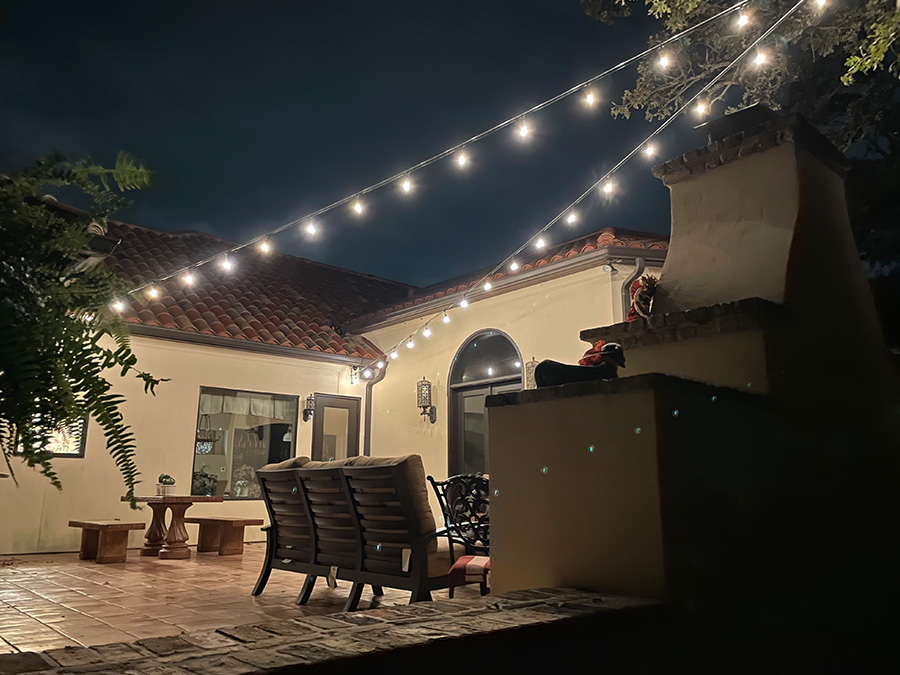 Professionally installed outdoor patio lights. Two strings of lights go over an outdoor kitchen with three chairs, a wood table with benches. The lights accent the stucco walls and tile roof.