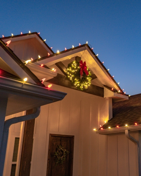 Font porch of a home with white and red holiday lights on the roof. A Christmas wreath wrapped in lights with a bow hangs above the front porch.