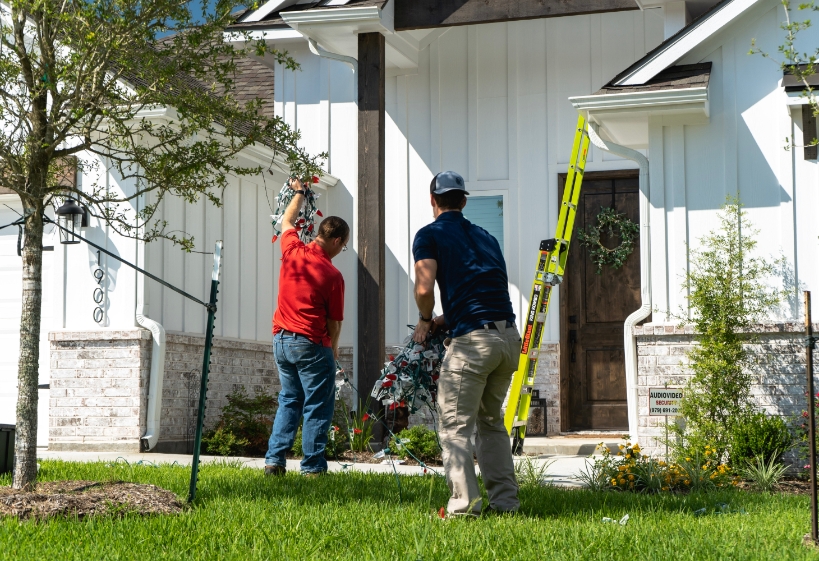 Two professional Christmas light installers unrolling red and white Christmas lights in front of a white farmhouse style home. There is a ladder against the roof next to the front door.