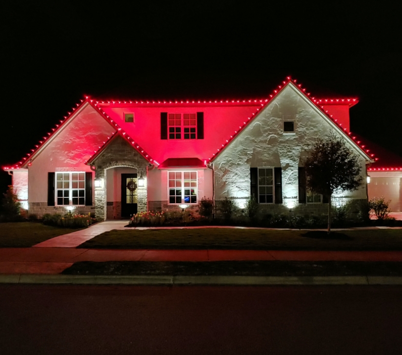 Perfectly spaced red Holiday lights line the roof of a white stone house. The house is glowing with red and white Christmas colors from the lights reflecting off of it.