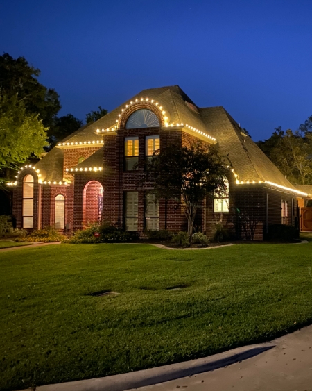 Red brick home in Bryan Texas with Christmas lights on the roof. The lights are reflecting off the roof making it look gold.