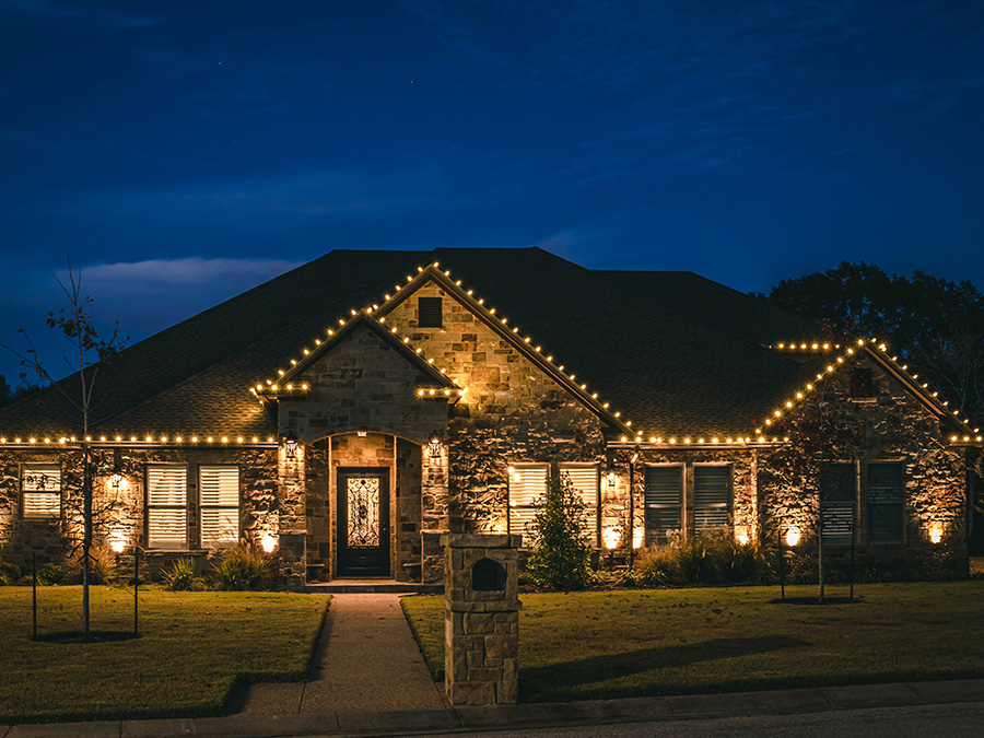 Warm Christmas lights installed on the front of a house. The beautiful landscape lighting and holiday lights show off the stone facade of the house.