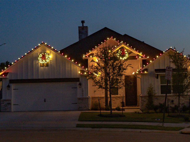 Holiday lights installed on a house in College Station TX. Lights line the roof, lit Christmas wreaths are installed over the garage and front door.