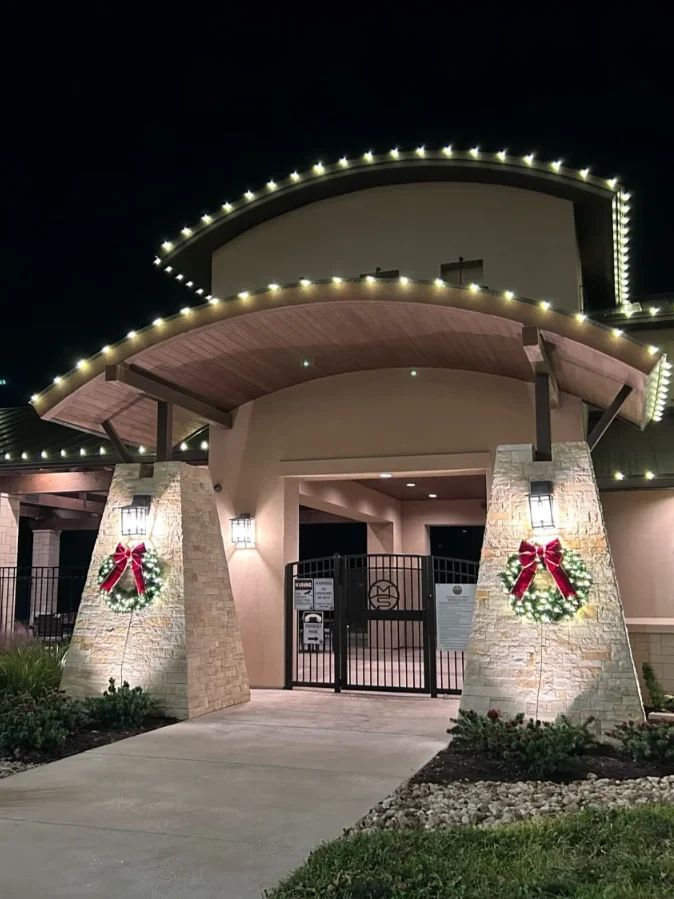 Front entrance of a business lined with Christmas lights, wreaths on pillars on each side of the door