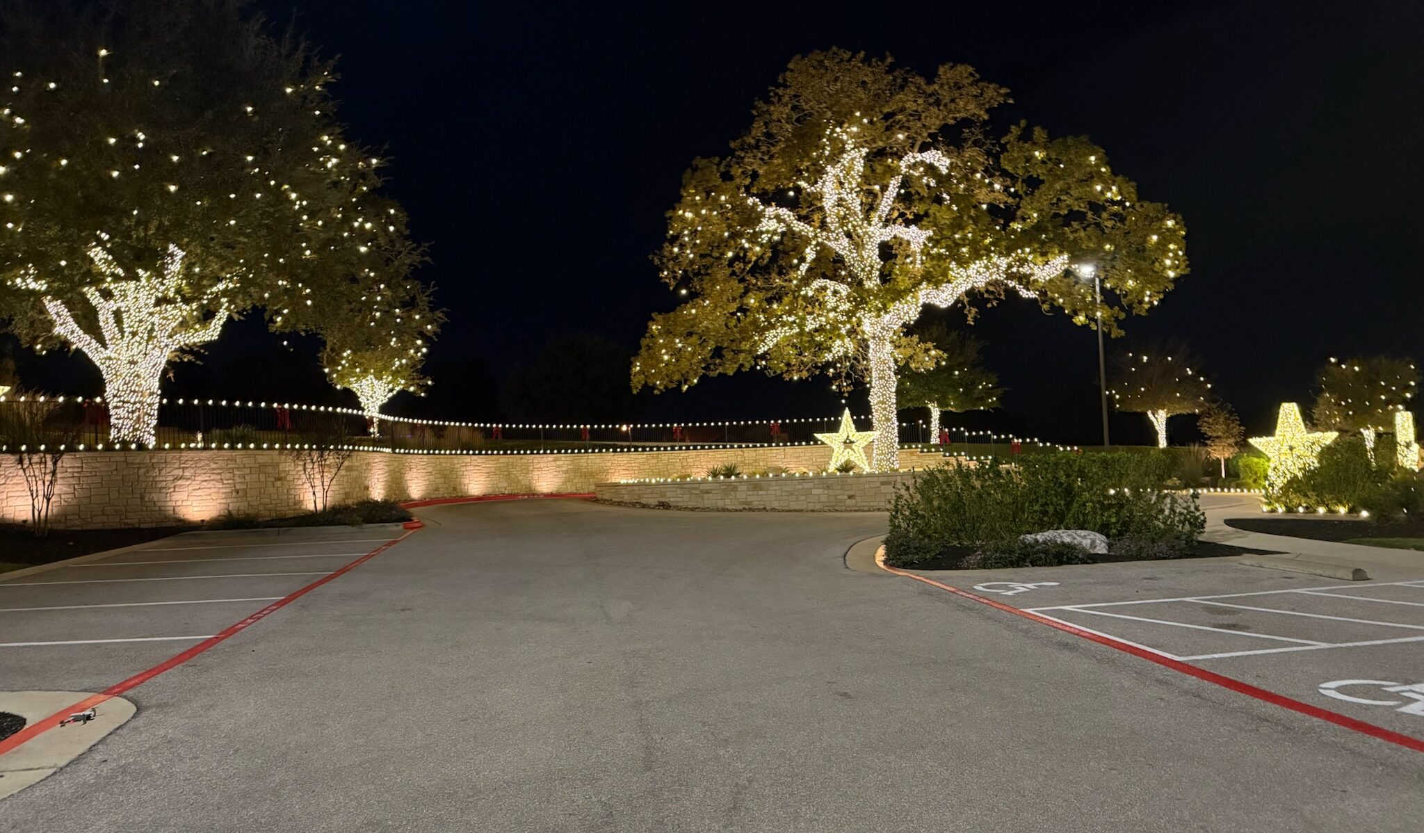 Neighborhood entrance with oak trees wrapped in warm white lights, a lit stone wall, and lighted stars